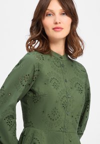 Green button-up dress with a textured pattern, featuring embroidered cut-outs. Long sleeves and a fitted bodice, flaring at the waist.