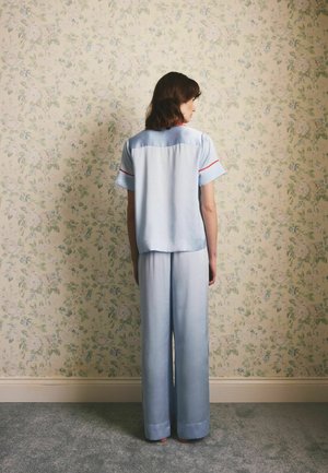 Žena stojaca bosá tvárou k kvetinovej tapete, oblečená v svetlomodrých voľných pyžamách s červeným lemom na rukávoch a golieri.