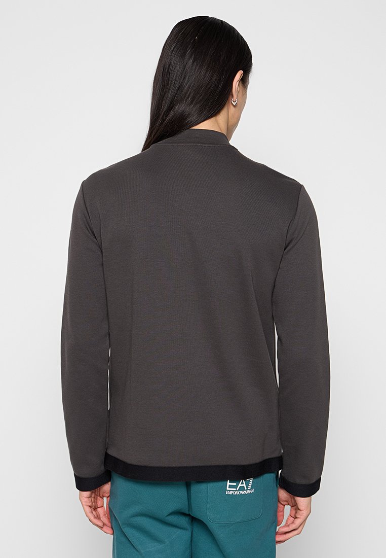 Maglietta grigia a maniche lunghe con colletto rotondo, texture a coste e orlo nero a bande. La vista posteriore mostra un design senza cuciture e dettagli minimi.