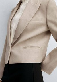 Blazer in lino beige con un design strutturato, revers a punta, dettaglio di una tasca singola e una silhouette accorciata, abbinato a una camicia di colore chiaro.