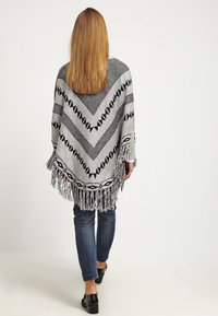 Kobieta z długimi blond włosami idąca w stronę z tyłu, ubrana w ponczo w czarno-białe wzory z frędzlami, ciemne dżinsy i czarne buty.