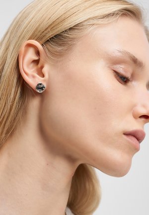 Nærbillede af en kvindes profil, der viser glat hud, blond hår der er stukket bag øret, og en lille, uregelmæssig sølvstuds.