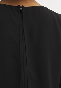 Musta mekko, jossa on sileä kangas, korkea kaula-aukko ja vetoketju keskiselässä. Suunnittelu on yksinkertainen, puhtain linjoin ja ilman koristeluja.