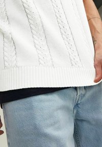 Weißer Strickpullover mit Kabelmustern, geripptem Saum und strukturierter Oberfläche, kombiniert mit hellblauen Jeans.