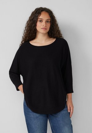 Zwarte oversized trui met een ronde halslijn en driekwart mouwen, gemaakt van een zachte stof. Gepaard met blauwe spijkerbroek.