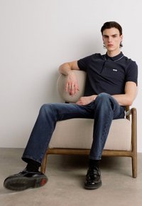 Marineblå polo med kontrasterende krave, slim-fit mørkeblå jeans, sorte lædersko, siddende i en beige polstret lænestol med træramme.