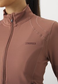 Brūns sporta jaka ar stāvošu apkakli, rāvējslēdzēju, izteiksmīgu "STRONGER" zīmolu, ar gludu audumu un dekoratīvo adījumu.