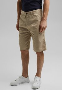 Khaaki szorty chino z płaskim przodem, bocznymi kieszeniami i zapięciem na guziki. Wykonane z lekkiej bawełnianej tkaniny. Połączone z białymi sneakersami.