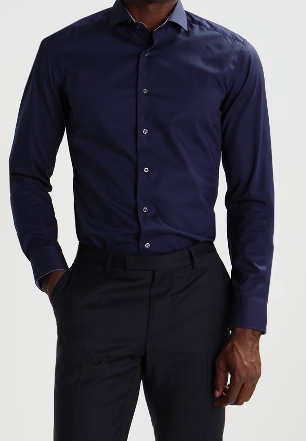 Homme portant une chemise cintrée bleu marine foncé à manches longues boutonnée, rentrée dans un pantalon de costume noir, debout avec une main dans la poche.