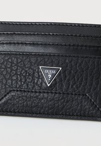 Mustmust nahkrahakott mitme kaardipesaga ja kolmnurkse hõbedase logoga, millel on "GUESS USA". Õmblemise detailid on nähtavad.