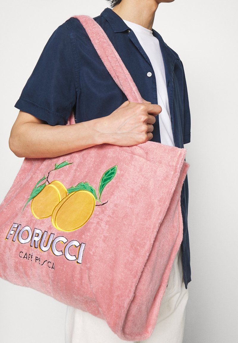 Personne en chemise bleu marine porte un grand sac Fiorucci rose avec des pêches jaunes et des feuilles vertes brodées, texte : "CAFFÈ PESCA."