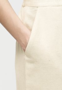 Χέρι που έχει εισαχθεί στην μπροστινή τσέπη μπεζ λινών παντελονιών, δείχνοντας την υφή του υφάσματος και τις λεπτομέρειες της ραφής.