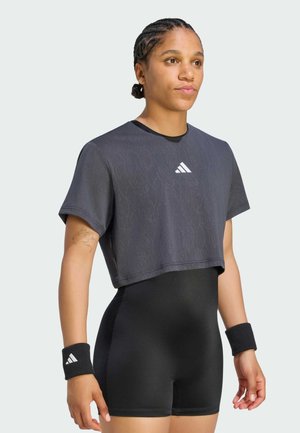 Młoda osoba z warkoczami, ubrana w ciemną krótką koszulkę Adidas, czarne dopasowane szorty i czarne opaski na nadgarstki z logo Adidas.