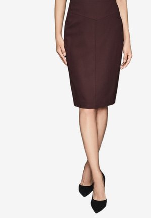 Femme portant une jupe crayon marron foncé ajustée longueur genoux et des talons aiguilles noirs pointus, debout les jambes croisées.