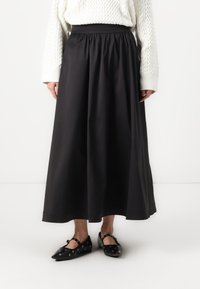 Black, ankle-length skirt with a gathered waistband and smooth texture, paired with flat black shoes featuring circular cutouts.