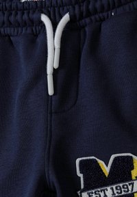 Morska trenerka z elastičnim pasom in belim vrvico. Ima teksturiran vezen logotip v rumeni in modri barvi. Mehka tkanina.