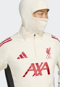 Person iført en krämfarvet Liverpool FC sportsjakke med røde logoer, herunder AXA og Adidas, samt en matchende ansigtsmaske.