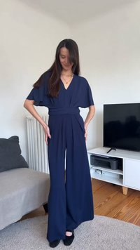Femeie cu păr lung și castaniu, purtând un combinezon bleumarin cu picioare largi și pantofi negri, stând într-un living cu televizor și canapea.