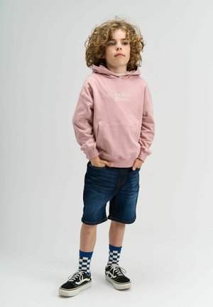 Hanorac roz deschis cu textul "Doar Cinci Minute În Plus", șorturi din denim bleumarin, adidași negri și șosete în carouri albastre. Păr creț.