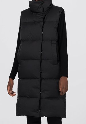 Czarna pikowana długa bezrękawnikowa kamizelka z wysokim kołnierzem, noszona na osobie na czarnej bluzce z długim rękawem, na jasnym tle.