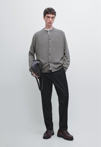 Cardigan gris clair avec un col rond, une fermeture à boutons sur le devant et des poignets côtelés, associé à un pantalon noir et des chaussures en cuir marron, tenant un sac.