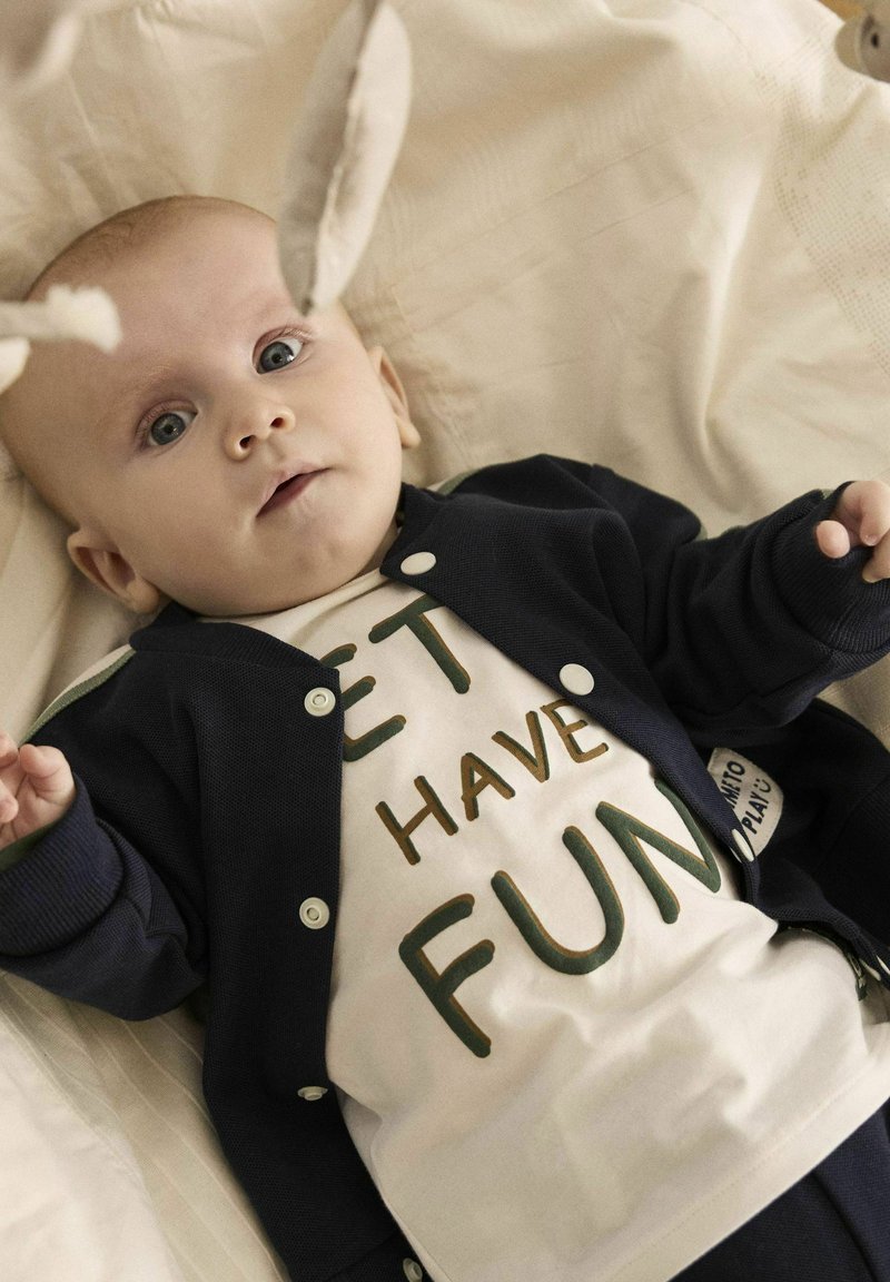 Marineblauer Cardigan mit weißen Druckknöpfen über einem cremefarbenen Hemd mit grünem Text "LASS UNS SPAß HABEN." Baby liegt auf einem hellbeigen Stoff.