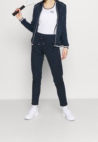 Mörkblå sportjacka med vita detaljer, tillsammans med matchande mörkblå joggingbyxor och vita sneakers; håller i en tenniskracket.