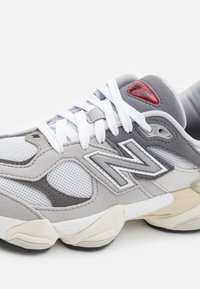 Grå og hvit joggesko med mesh- og semsket skinnmaterialer, en kraftig såle, fremtredende "N"-logo, og rød aksent nær hælen.