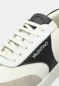 Baskets en cuir blanc avec des accents noirs et beiges, présentant des textures lisses et en daim, des lacets plats, et un logo sur le côté.