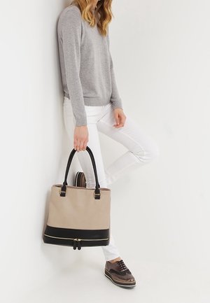 Femme appuyée contre un mur, portant un pull gris, un pantalon blanc, des chaussures brogues marron, tenant un sac à main beige et noir avec deux poignées.