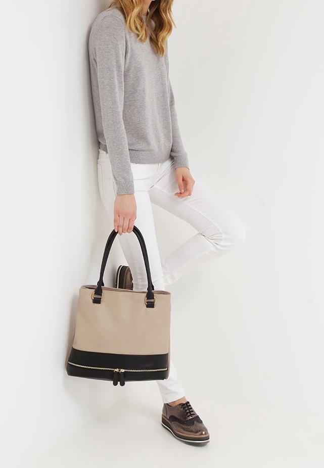 Femme appuyée contre un mur, portant un pull gris, un pantalon blanc, des chaussures brogues marron, tenant un sac à main beige et noir avec deux poignées.