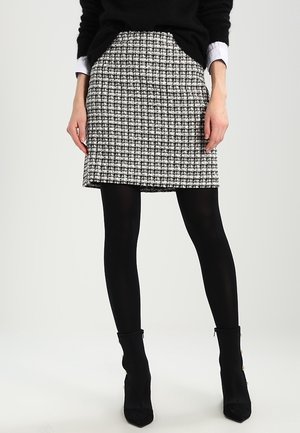 Femme portant une jupe à carreaux noir et blanc, des collants noirs, des bottines noires et un pull noir avec des poignets de chemise blancs visibles.