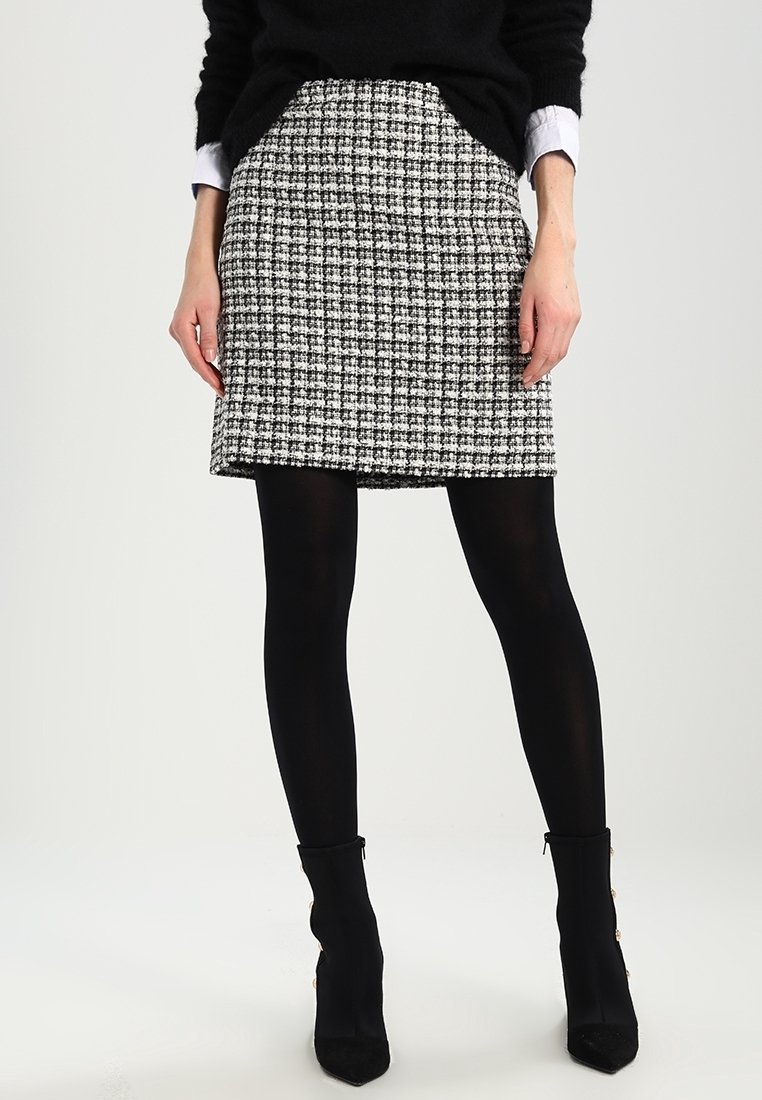 Femme portant une jupe à carreaux noir et blanc, des collants noirs, des bottines noires et un pull noir avec des poignets de chemise blancs visibles.