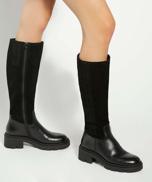 Bottes noires montantes jusqu'au genou avec semelle épaisse et fermeture éclair sur le côté, portées sur des jambes nues sur un fond uni.