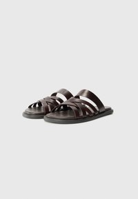 Des sandales à glisser en cuir marron présentent un design à lanières entrecroisées, une semelle en caoutchouc plate et une forme minimaliste avec des bouts ouverts.