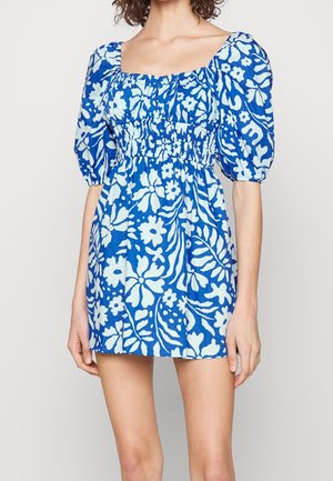 Kobieta ubrana w niebiesko-białą mini sukienkę w kwiaty, z bufkami na krótkich rękawach i marszczonym kwadratowym dekoltem.