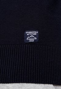 Marintröja i blått, stickad med ribbat avslut. Har en fyrkantig tyg-etikett med "Springfield Attitude" och bergdesign, daterad 1988.