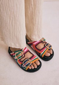 Farverige vævede sandaler med dobbelt spænde, en tyk sort sål og et flerfarvet stribet mønster i pink, grøn og orange.