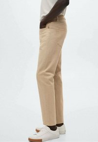 Chinos de algodón beige con corte recto, dos bolsillos laterales y una cinturilla clásica. Llevados con zapatillas blancas. Textura suave.
