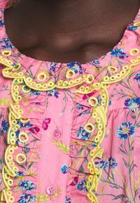 Rosa blus med blommönster som har gul broderad kant och volangkrage. Inkluderar knappar och botaniska mönster i blått och grönt.