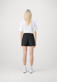 Melni šorti ar augstu jostasvietu un divām aizmugurējām kabatām, apvienoti ar baltu īsu t-kreklu. Modelis ir pagriezies ar muguru, valkājot baltus sporta apavus.