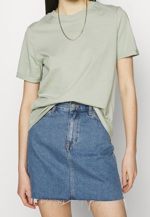 Donna che indossa una t-shirt verde chiaro a maniche corte infilata in una mini gonna di jeans blu con orlo sfrangiato.