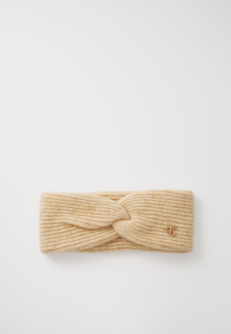 Knit headband in light beige, featuring a twisted design at the front and subtle ribbing. Small gold logo accent on the side.