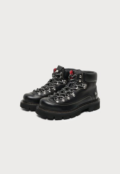 Paire de bottes de randonnée en cuir noir avec semelles épaisses, œillets métalliques, lacets noirs et blancs, et collets de cheville rembourrés sur un fond uni.