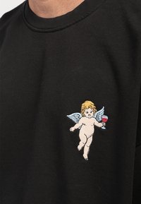 Črna športna majica z vezenim motivom cheruba, ki drži rdečo skodelico. Cherub ima blond lase in modre krila.