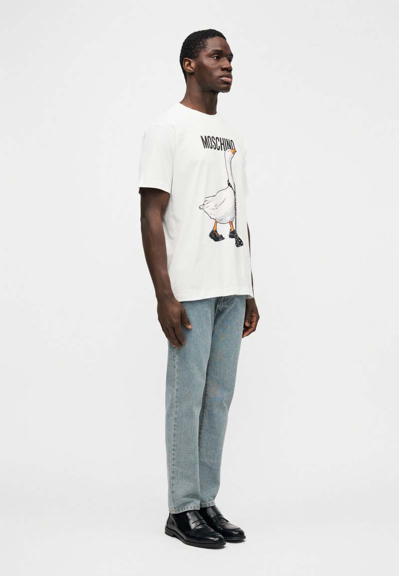 Weißes T-Shirt mit einer Grafik einer Ente, die schwarze Schuhe trägt, darüber mit der Aufschrift "MOSCHINO". Kombiniert mit hellblauen Jeans und schwarzen Schuhen.