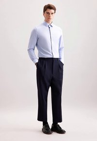 Camicia azzurra con bottoni, pantaloni plissettati blu scuro e scarpe nere. La camicia ha un colletto classico e maniche aderenti; i pantaloni sono a taglio dritto.
