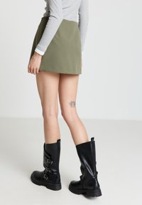 Olivgrüner Minirock mit glatter Textur und figurbetontem Schnitt, kombiniert mit kniehohen schwarzen Lederstiefeln mit Schnallenakzenten.