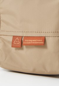 Beige recycled nylon bag with textured surface. Features a brand tag and a label stating, “This bag was trash!” in orange.