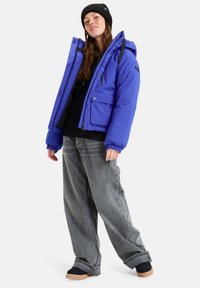 Blaues Pufferjackett mit Kapuze, elastischen Bündchen und Fronttaschen, kombiniert mit lockeren grauen Jeans und schwarzen Schuhen. Accessoire: schwarze Mütze.